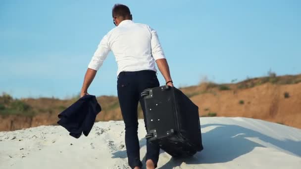 Бизнесмен в пустыне. Молодой человек в деловом костюме и большая сумка для багажа в пустыне. Бизнесмен на пути к курорту. Белый песок. Знойная пустыня . — стоковое видео
