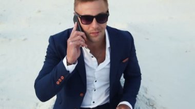 İşadamı çölde çalışma ile bir dizüstü bilgisayar ve cep telefonuyla konuşuyor. Güneş gözlüğü ve bir takım elbise genç adam.
