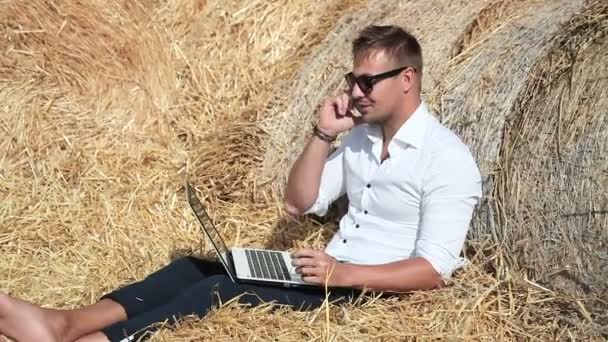 Деловой человек выглядит прекрасно работает с ноутбуком и разговаривает по телефону, сидя в стоге сена. Летние каникулы за городом. Работа откуда угодно. Фрилансер на работе . — стоковое видео