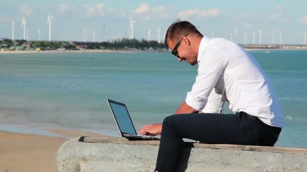 Obchodní muž v bílé košili a sluneční brýle s notebookem u pobřeží. V pozadí, moře, zarděnky, kuřecí turbíny elektrárny. Pracujte kdekoli na světě. Na volné noze v práci. — Stock video
