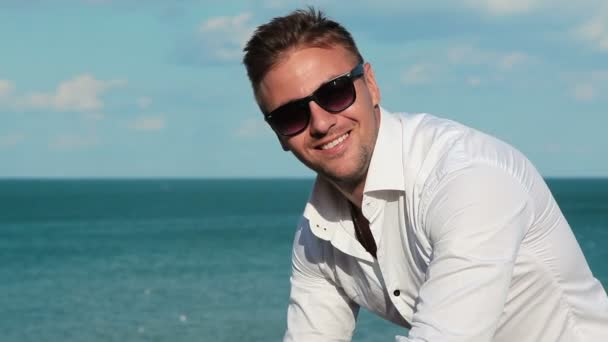 Hombre de negocios con camisa blanca y gafas de sol con un portátil en la orilla del mar. En el fondo, el mar, la rubéola, las plantas de energía de turbina de pollo. Trabaja desde cualquier parte del mundo. Independiente en el trabajo . — Vídeos de Stock