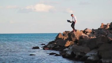 İş adamı bir dizüstü bilgisayar ile zafer sevinci ile deniz kenarında büyük kayalar üzerinde elini yukarı kaldırır. Zafer sevinci. Başarı elde etmek.