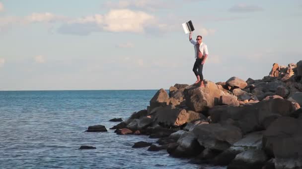 Hombre de negocios en grandes rocas junto al mar con la alegría de la victoria con un portátil levanta la mano. La alegría de la victoria. Lograr el éxito . — Vídeos de Stock