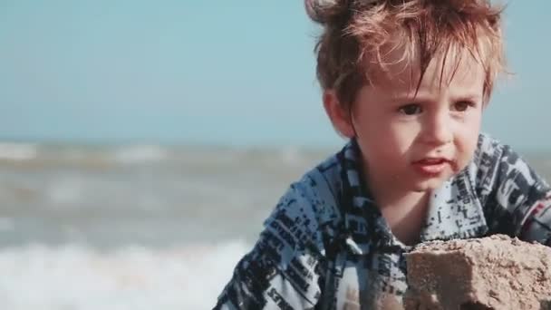 Pojke 2 år gamla, vackra utseende, spelar i sanden på stranden. De våta kläderna och kul. Sommardag vid havet. — Stockvideo