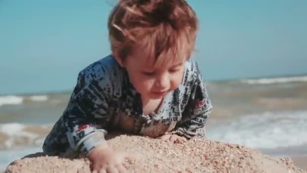 Jongen leeftijd 2 jaar oude, mooie verschijning, spelen in het zand aan de kust. De natte kleren en plezier. Zomerdag aan zee. — Stockvideo