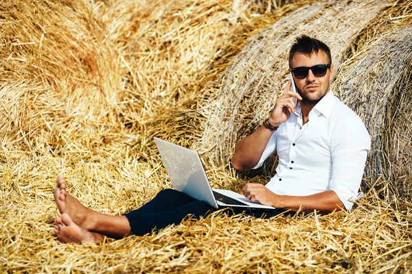 Άνθρωπος των επιχειρήσεων φαίνεται όμορφα έργα με ένα φορητό υπολογιστή και μιλάει στο τηλέφωνο κάθεται σε άχυρα. — Φωτογραφία Αρχείου