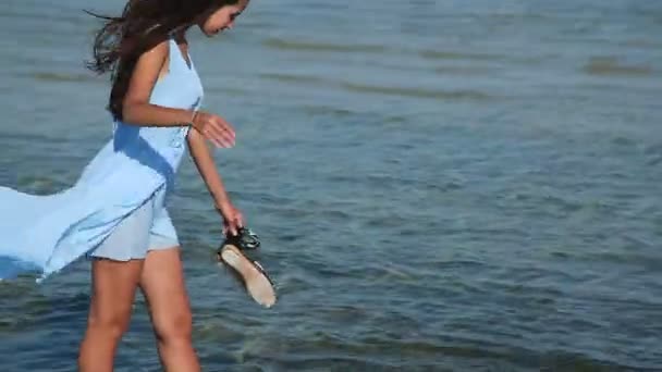 Όμορφο και ελκυστικό κορίτσι ήρθε στην παραλία και πήγε μέσα στο νερό. Το κορίτσι με τα πόδια στην παραλία. Κορίτσι με ένα μπλε φόρεμα, και πήγε στη θάλασσα. Πιτσιλίσματα νερού. — Αρχείο Βίντεο