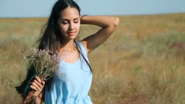 Menina bonita nova em um vestido azul gosta de flores estepe. Uma jovem mulher num prado. Estepes. Prado ensolarado florescente com grama alta espessa orelhas e flores — Vídeo de Stock