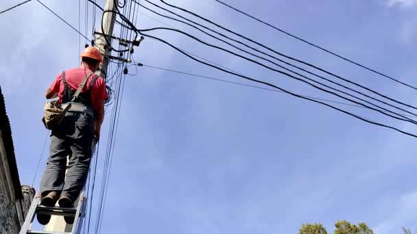 Электрик в шлеме чинит провода на лестнице возле высокого полюса — стоковое видео