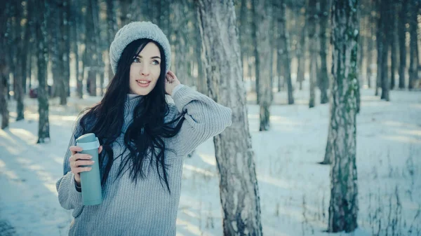 Elinde termo bardakla çam ormanında süveter ve şapka giyen genç bir kadın..