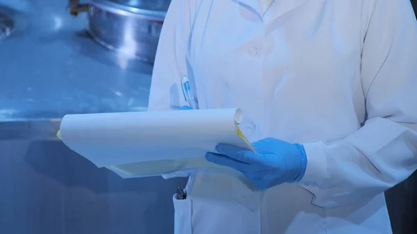 Bir kadın, bir üretim atölyesinde, beyaz önlüklü, steril eldivenli belgeleri elinde tutuyor.