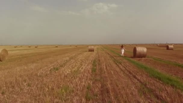 Жінка в білій спідниці біжить уздовж поля з прокатом соломи — стокове відео