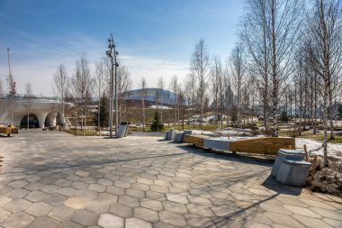 Zaryadye Park manzarası ilkbaharın başlarında güneşli bir gün: bir huş ağacında ahşap banklar ile Amphitheater ve Kotelnicheskaya Embankment Binası karşı.