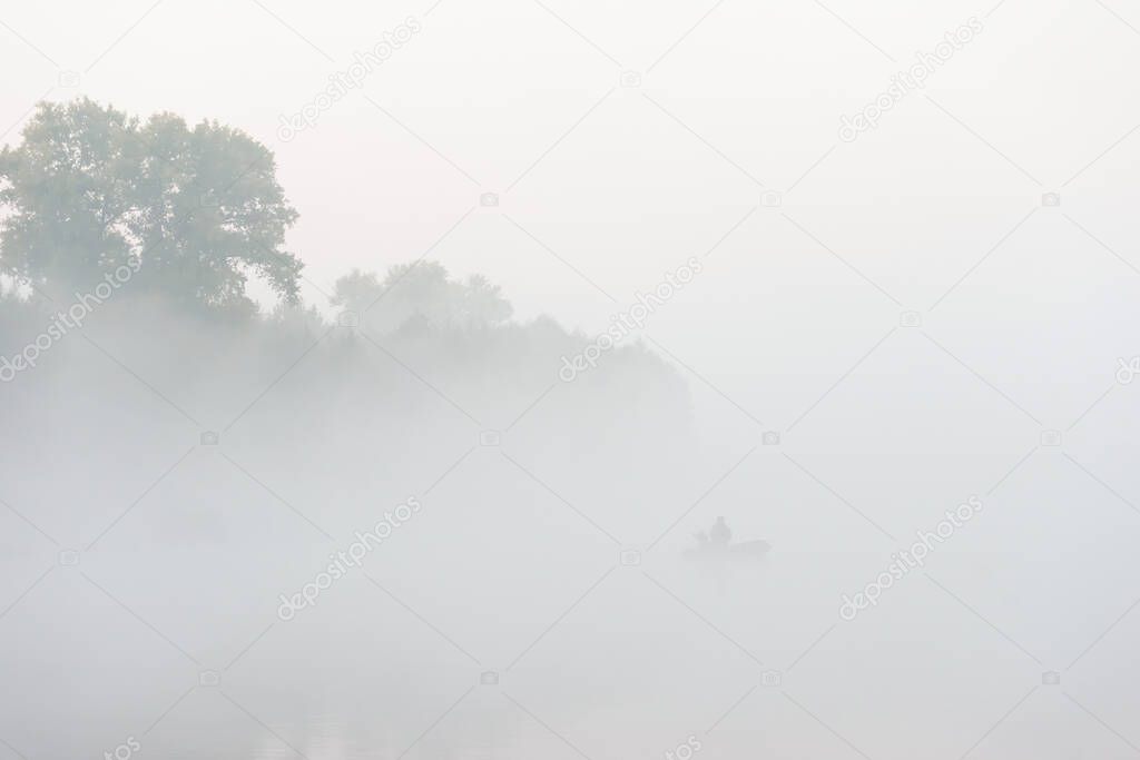 Silhouette of fisherman sitting in rubber boat on a river at misty morning