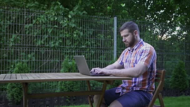 Man schrijven op een laptop, zittend op houten stoel in kleine tuin. — Stockvideo