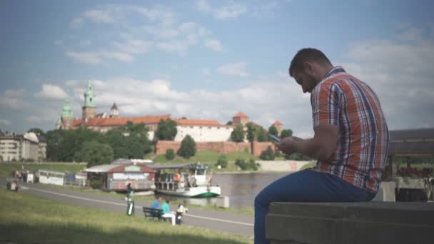 Чоловік за допомогою смартфона сидить на стіні біля річки — стокове відео