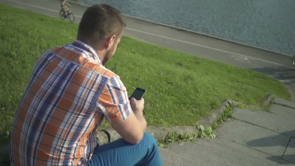 Man browsing smartphone, sitting on stairs by the river. — Stock Video