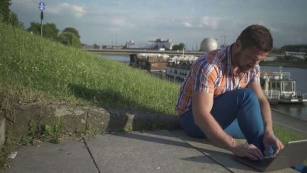 Man writing on a laptop on the stairs by the river, pan shot, right. — Stock Video