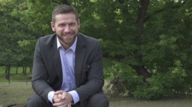 Parkta oturan kameraya gülümseyen iş adamı.