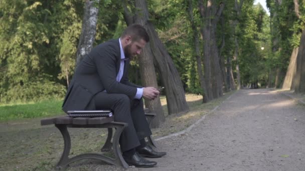 Busienessman 공원에서 나무 벤치에 앉아 스마트폰 검색, 팬 촬영. — 비디오