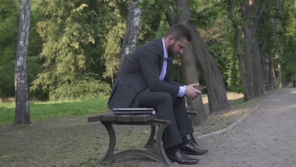 Busienessman naviguant sur le smartphone assis sur un banc en bois dans le parc, pan shot . — Video