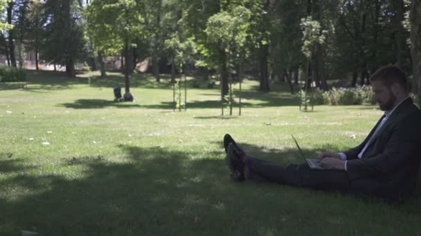 Empresario sentado bajo el árbol en el parque, trabajando en el portátil. Pan Shot . — Vídeo de stock