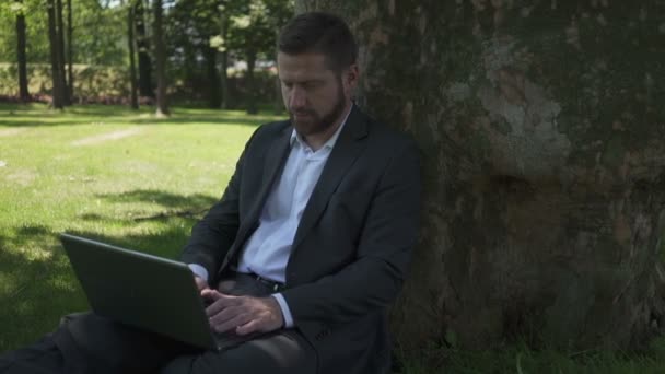 Geschäftsmann sitzt unter einem Baum im Park und arbeitet am Laptop. Schwenk geschossen. — Stockvideo