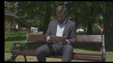 İşadamı, parkta ahşap bankta otururken tabletle selfie çekiyor.