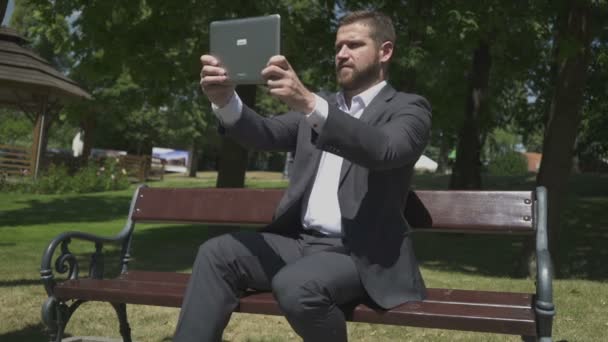 Επιχειρηματίας παίρνει πανοραμική φωτογραφία με δισκίο, καθισμένος σε ξύλινο παγκάκι στο πάρκο. — Αρχείο Βίντεο