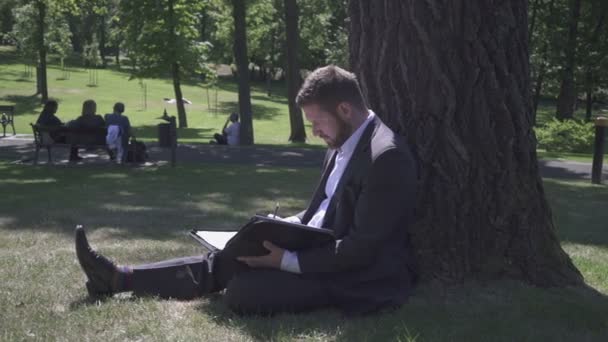 사업가는 공원에서 나무 아래에 앉아, 문서와 함께 작업하고 있습니다. 팬 샷. — 비디오