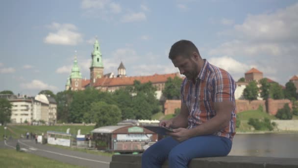 Young man browsing tablet sitting on wall. — Stock Video