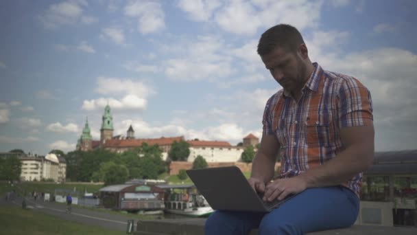 Молодой человек печатает на компьютере, сидит на стене у реки . — стоковое видео