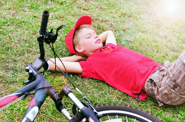 Boy in red T-shirt and cap is sleeping on the green grass with his bicycle.