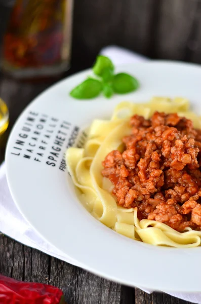 Pasta Bolognese in the white plate with letters, with basil leaf. Little bowl of olive oil and chili pepper on the white napkin on the old wooden table — Stock Photo, Image