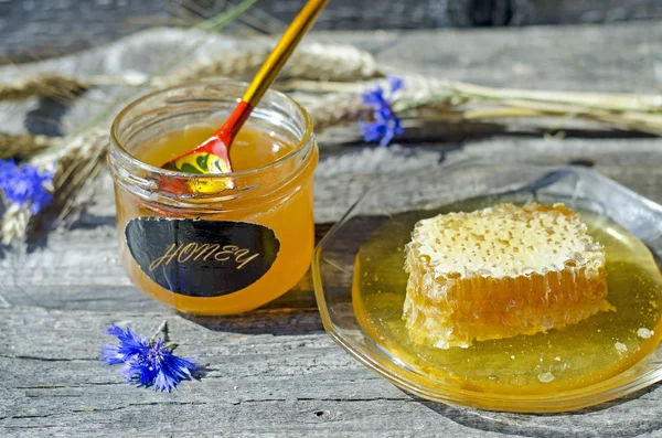 Honning i honeycomb på glaspladen og lille krukke med russisk ske med ører og kornblomster på træets baggrund, selektivt fokus - Stock-foto