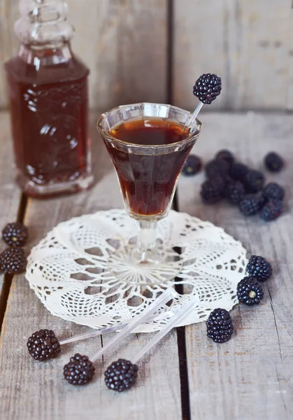 Blackberry liquor in the little glass and vintage bottle on the vintage napkin on the old wooden background, toned picture, selective picture — Stock Photo, Image
