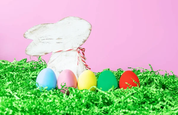 Fond pour Pâques avec une figurine d'un lapin avec des œufs colorés dans l'herbe sur un fond rose, endroit pour le texte, symbole de Pâques lumière — Photo