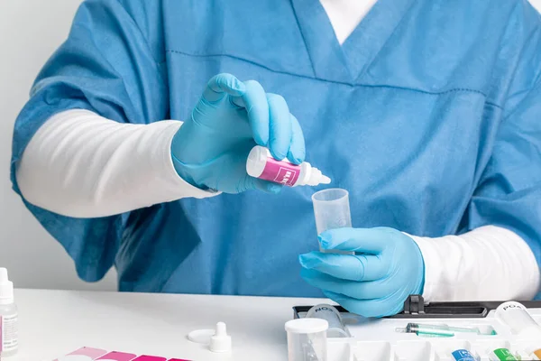 Tıbbi kıyafetli ve eldivenli bir laboratuvar asistanı suyu kimyasal reaktörlerle test eder.