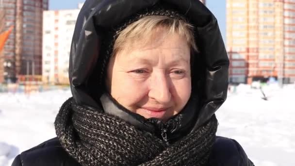 Old middle-aged woman stands in the street in winter looking at the camera, smiles, laughs, in a black down jacket hood, a scarf against the background of residential buildings on a sunny day, close — Stock Video