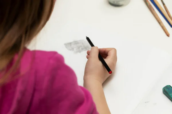 Little girl sits at a white table, holds a pencil in her hand and is engaged in drawing, back view, lifestyle — Stock Photo, Image