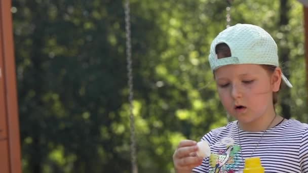 Маленькая девочка в шапке и полосатой футболке надувает мыльные пузыри на улице на фоне деревьев в солнечный день, копирует пространство — стоковое видео