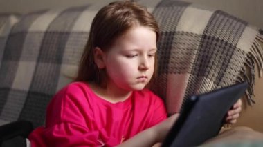 Küçük, sevimli, uzun saçlı güzel bir kız oturur ve oynar, rahat bir odada tablet kullanır, ekoseli bir hayat tarzının arka planına karşı.