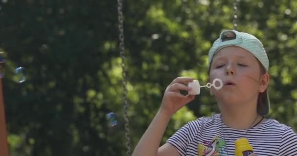 Маленька мила дівчинка в шапці і смугаста футболка грає з мильними бульбашками, дме їх на дитячий майданчик в сонячний літній день — стокове відео