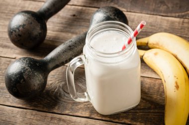 Beyaz protein kokteyli, klasik halterler ve ahşap masada muz meyvesi.