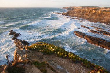 Kayalıklarla kaplı engebeli kıyı manzarası, sörf dalgaları, denizdeki kayalar, yumuşak güneş ışığıyla aydınlatılmış.