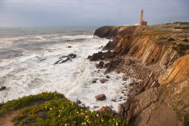 Kayalıklarla kaplı engebeli kıyı manzarası, sörf dalgaları, denizdeki kayalar, yumuşak güneş ışığıyla aydınlatılmış.