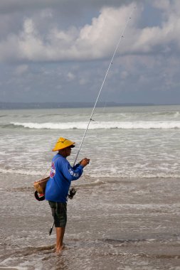 Sahilde şapkalı bir adam balık tutuyor.