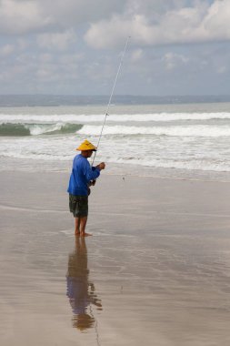 Sahilde şapkalı bir adam balık tutuyor.