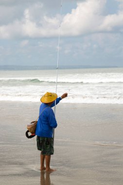 Sahilde şapkalı bir adam balık tutuyor.
