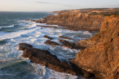 Kayalıklarla kaplı engebeli kıyı manzarası, sörf dalgaları, denizdeki kayalar, yumuşak güneş ışığıyla aydınlatılmış.